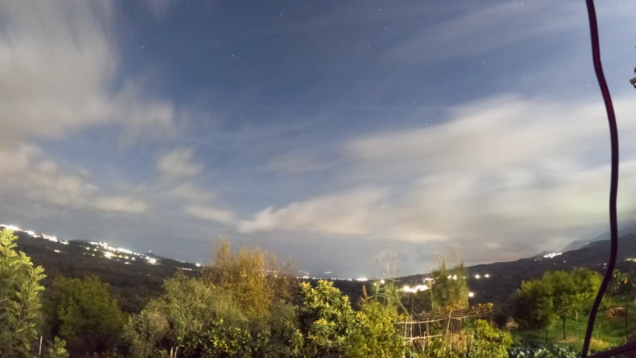 Time lapse 4K del cielo notturno con nuvole in rapido movimento e stelle sopra paesaggi rurali e luci della città