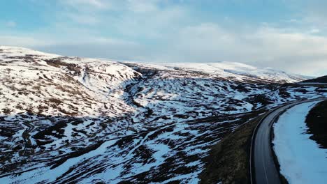 Aerial-view-of-winter-landscape-with-asphalt-road-with-sunlight-falling-on-snow-covered-mountains,-Iceland