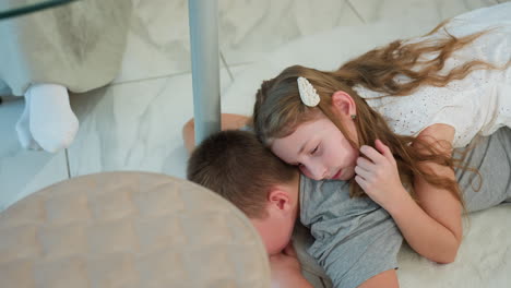 girl in white gown relaxing comfortably on brother s back, enjoying playful moment, with view of woman s leg in white socks under table, cozy indoor scene with family, sibling bonding time