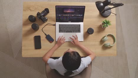 top view of asian woman color grading about the footage then raising index finger while sitting in the workspace using a laptop next to the camera editing the video at home