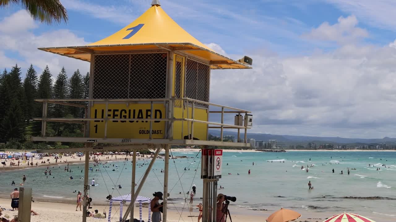 A yellow lifeguard tower stands watch over a crowded beach with swimmers and beachgoers.
