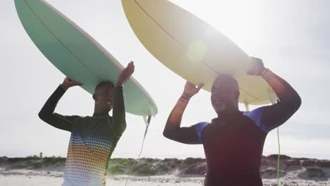 Afroamerikanischer-Vater-Und-Sohn-Im-Teenageralter-Stehen-Am-Strand,-Halten-Surfbretter-Auf-Dem-Kopf-Und-Reden