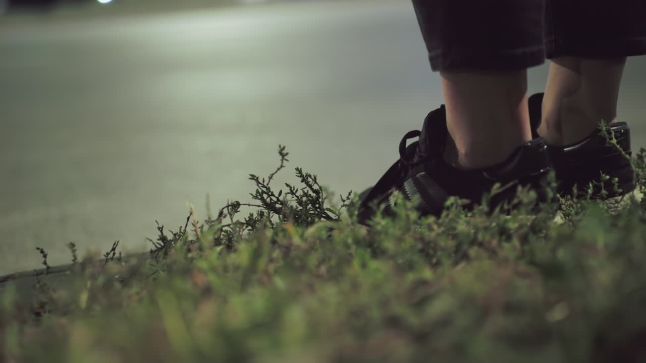 Leg view of person wearing black sneakers and cropped pants standing near roadside with green grass in foreground and blurred headlights of passing vehicles in distant background