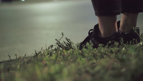 leg view of person wearing black sneakers and cropped pants standing near roadside with green grass in foreground and blurred headlights of passing vehicles in distant background