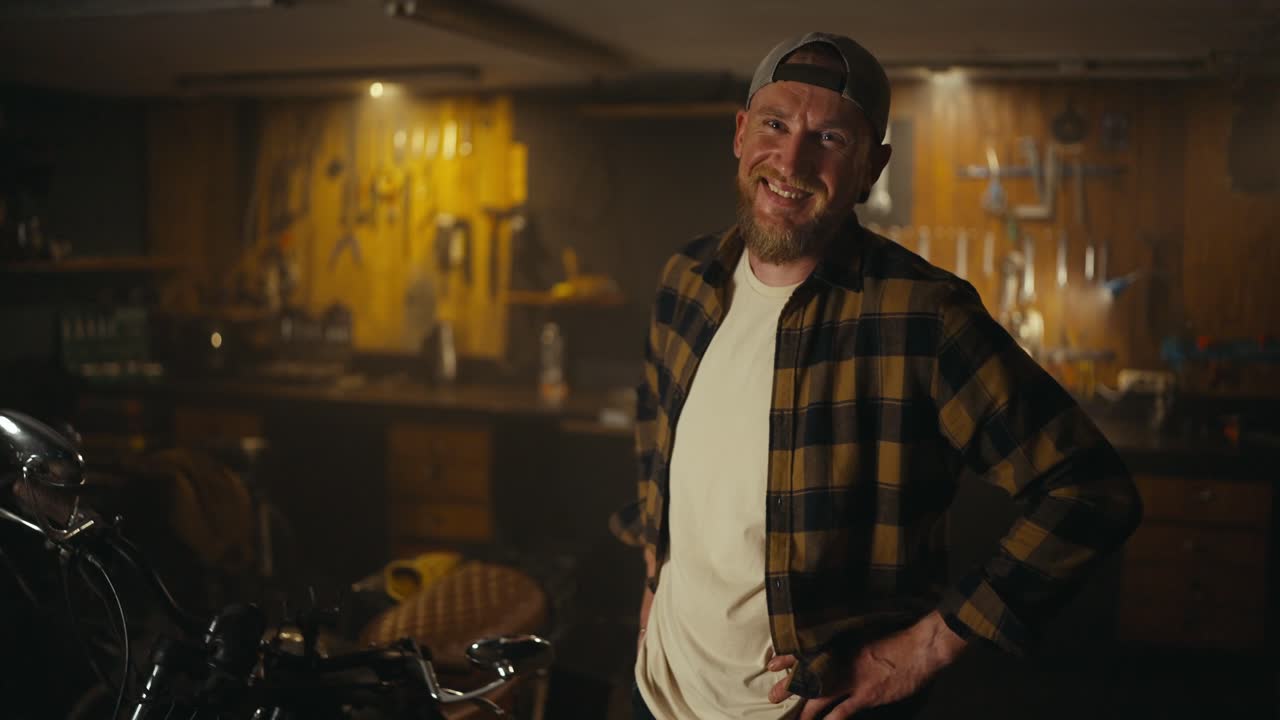 Portrait of a cheerful guy mechanic in a checkered shirt and cap posing and laughing in his workshop studio