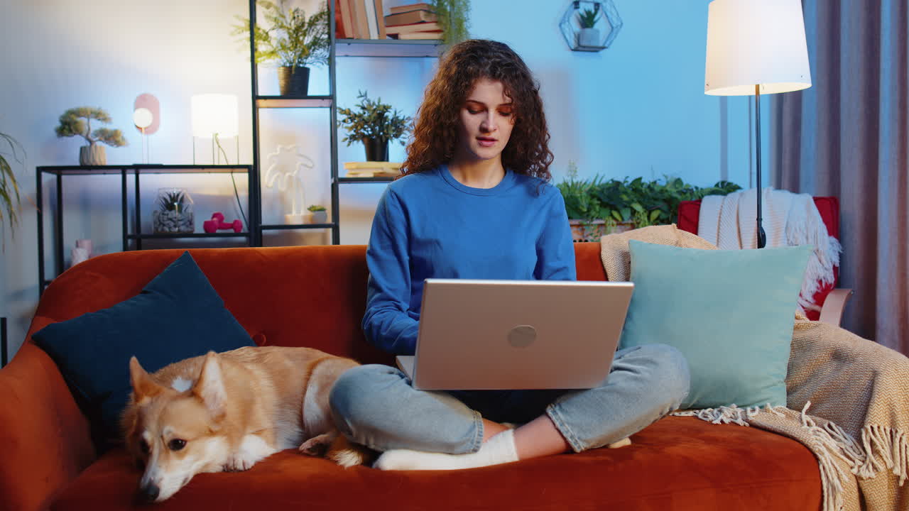 Smiling woman closing laptop after finishing remote work sitting beside corgi dog on home sofa