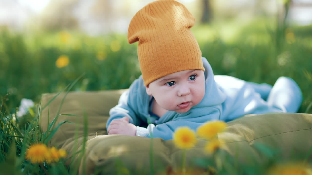 Healthy Caucasian baby boy lying on his belly on big pillow. Cute kid in orange cap resting in green grass. Blurred backdrop.