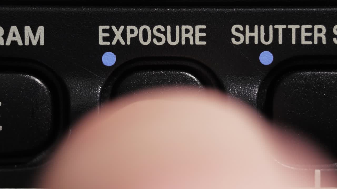 Close-up macro de um dedo pressionando o botão de controle de exposição com um ponto azul e letras brancas.