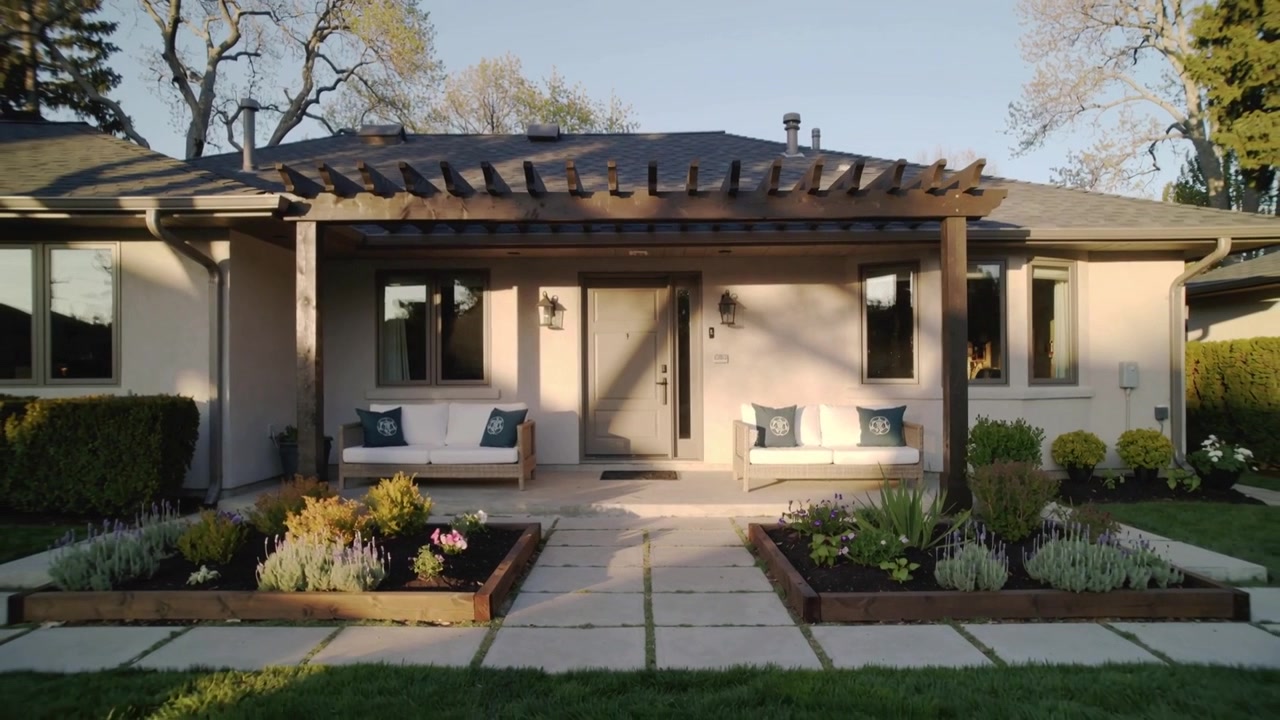 Telecamera che inizia a muoversi lentamente verso il portico di una casa a un piano, mostrando pergola, porta, panchine e fioriere.