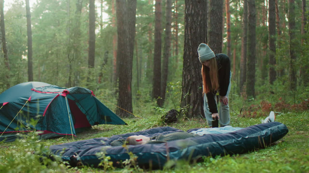Husband lays on tent bag while wife slightly bent pumps air into it, both smiling and gazing warmly at each other with tent behind them in peaceful forest campsite during outdoor bonding moment