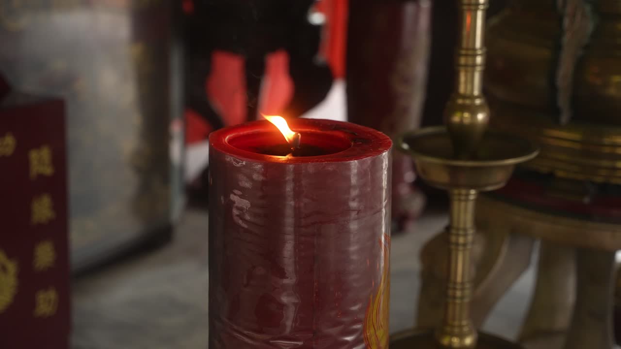 A close-up holds on the red candle as its flame burns bright. The fixed angle captures brass ceremonial objects behind, contrasting the vivid glow with intricate, shadowed cultural detail