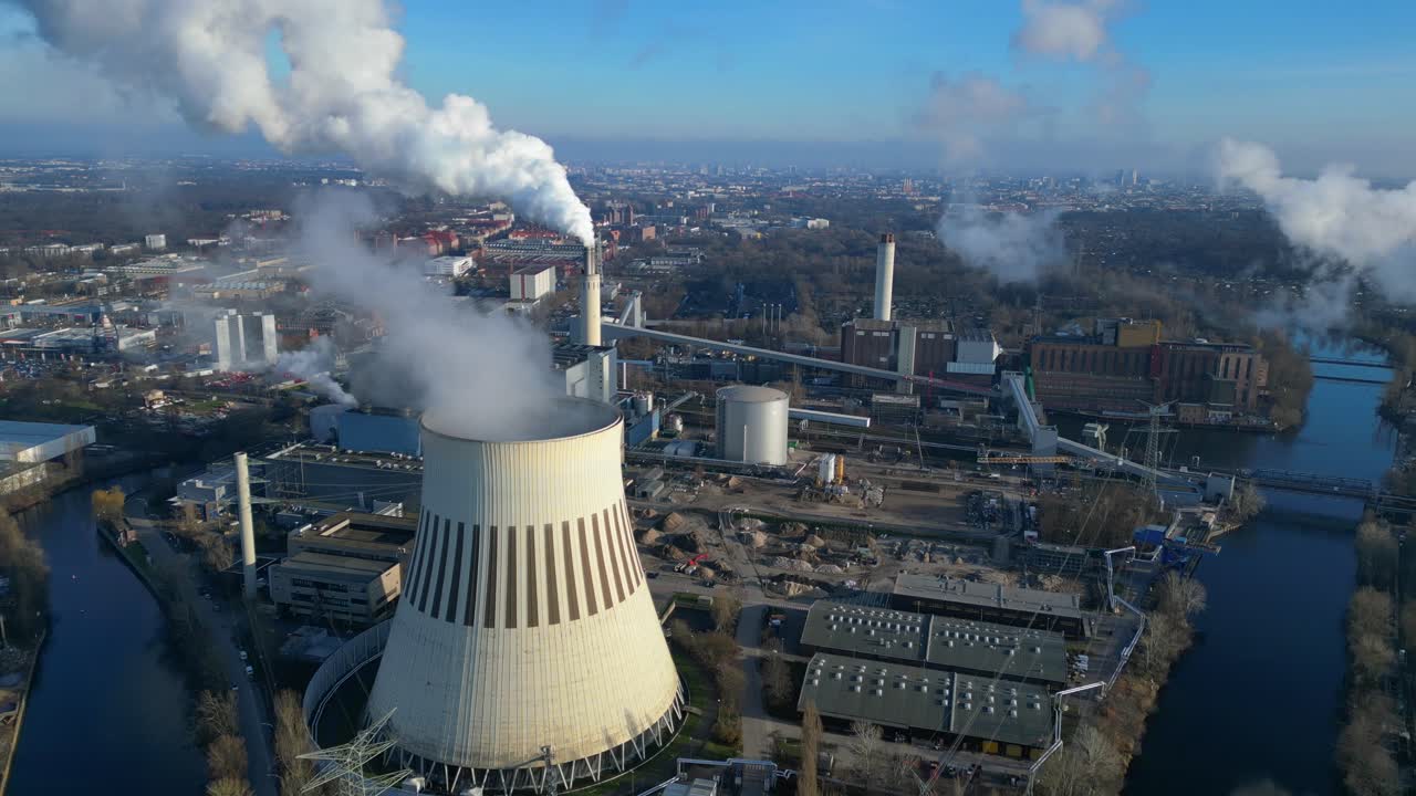 Aerial view of a thermal power plant emitting smoke on a sunny day, with a river and a city in the background. Best aerial view flight drone shot footage from above