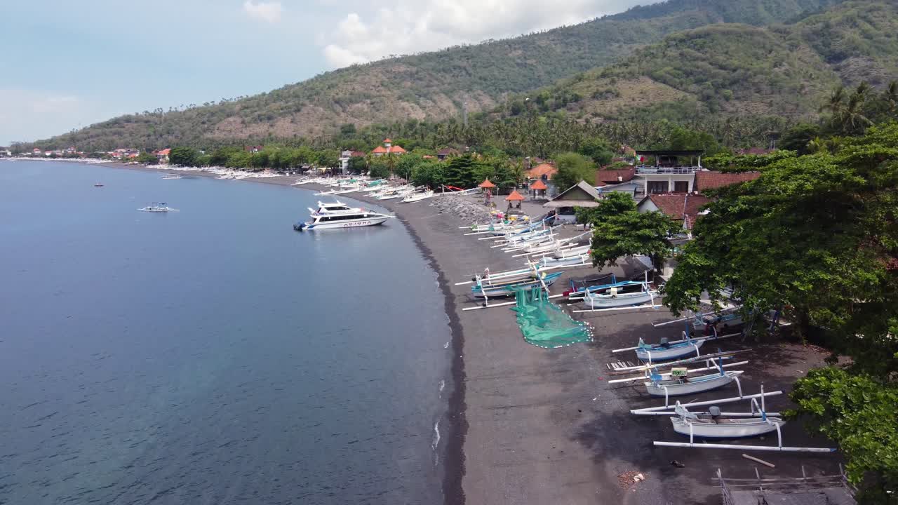 vista aérea de la costa del pueblo de amed con un yate amarrado en la bahía y barcos de pesca jukung blancos locales en la playa de arena volcánica negra, casas de los aldeanos y montañas en el fondo