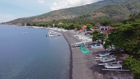 Vista-Aérea-De-La-Playa-Del-Pueblo-De-Amed-Con-Un-Yate-Amarrado-En-La-Bahía-Y-Botes-De-Pesca-De-Jukung-Blancos-Locales-En-La-Playa-De-Arena-Volcánica-Negra,-Casas-De-Aldeanos-Y-Montañas-En-El-Fondo