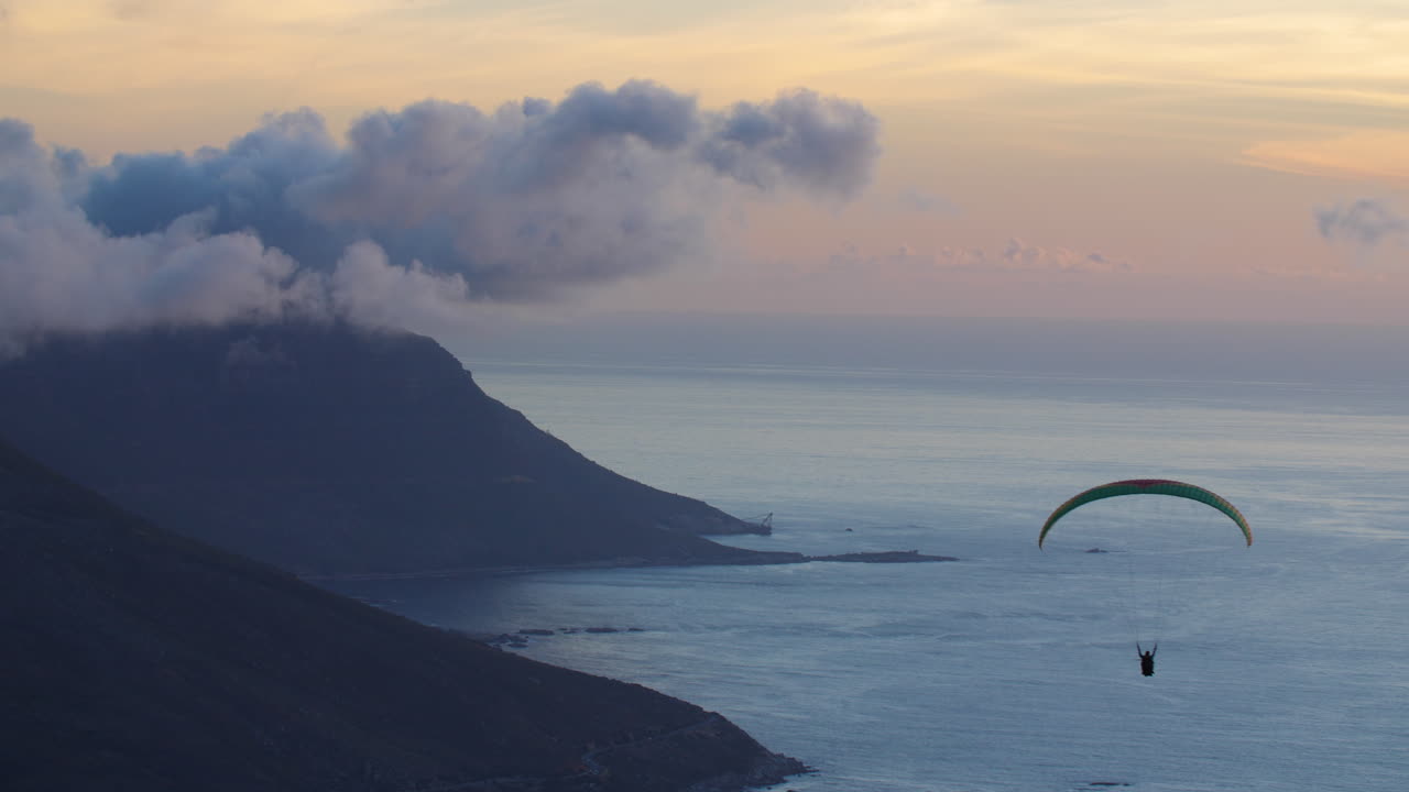 Paraglider flying over ocean along cloudy mountainous coastline at dusk