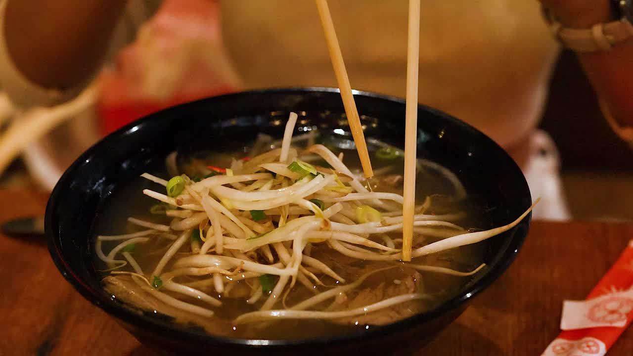 A person uses chopsticks to eat Vietnamese pho, highlighting the dish's ingredients and dining experience in warm lighting