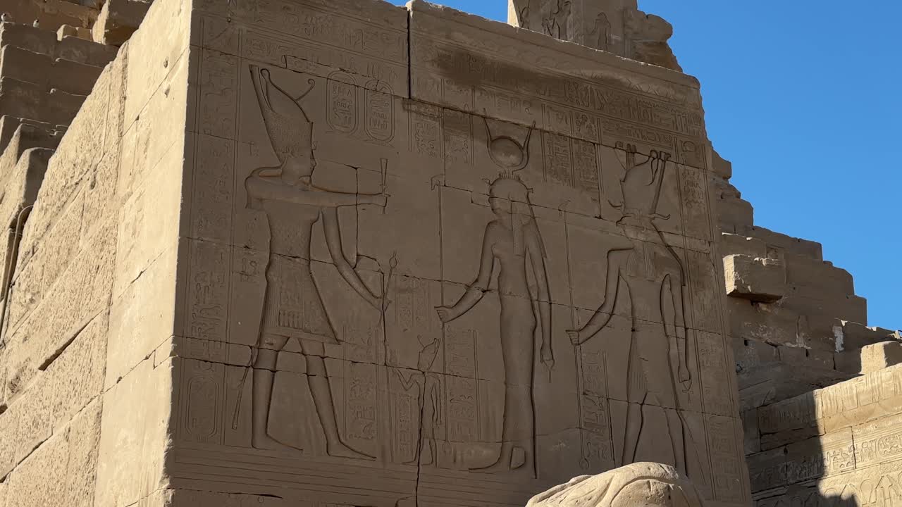Lähikuva hyvin säilyneestä muinaisesta egyptiläisestä reliefistä, joka esittää kolmea hahmoa ja hieroglyfejä monumentaalisessa kiviblokissa kirkkaan sinisen taivaan alla Denderan temppelissä.