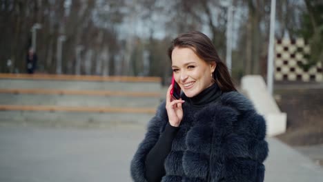 smiling woman talking the phone. cheerful girl in fur coat standing in the park and speaks on the mobile phone. slow motion.