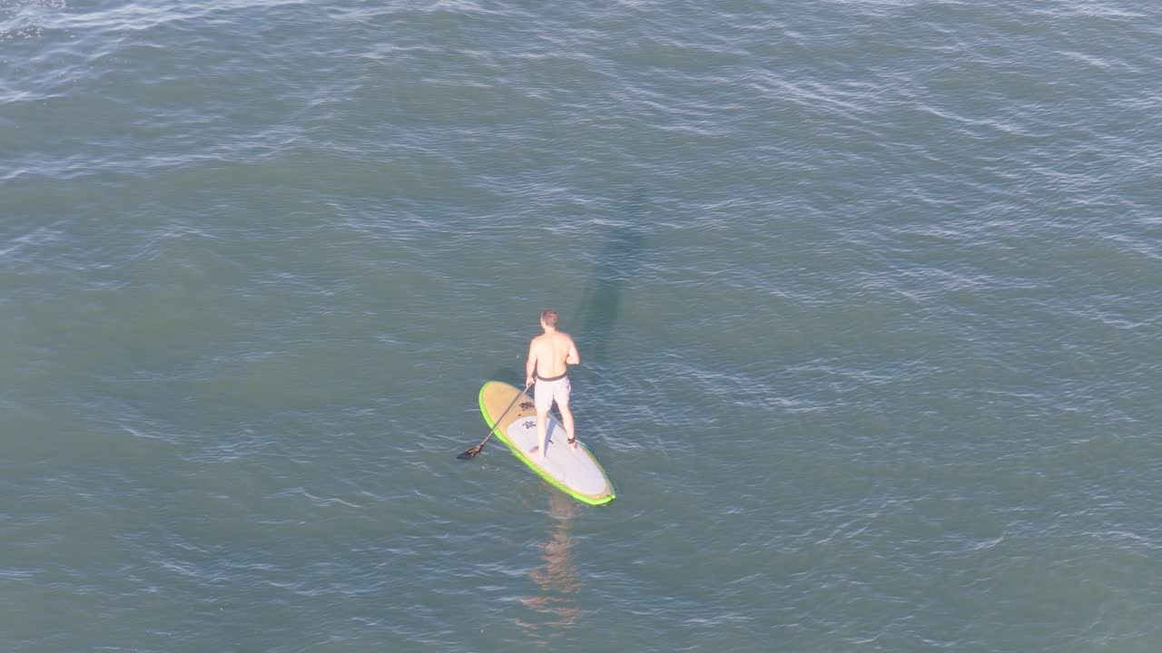Drone captures a man paddleboarding on calm ocean waters, highlighting serene outdoor activity and natural beauty
