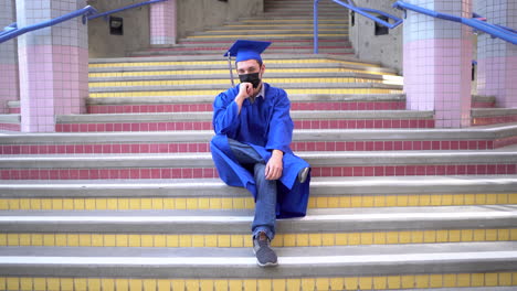 male graduate sits at school in his cap and gown with face-mask on