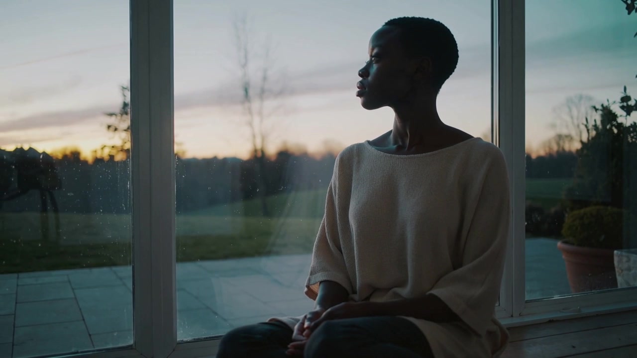 Sitting woman wearing light sweater, dark pants on window ledge, sunset drawing gaze, copy space