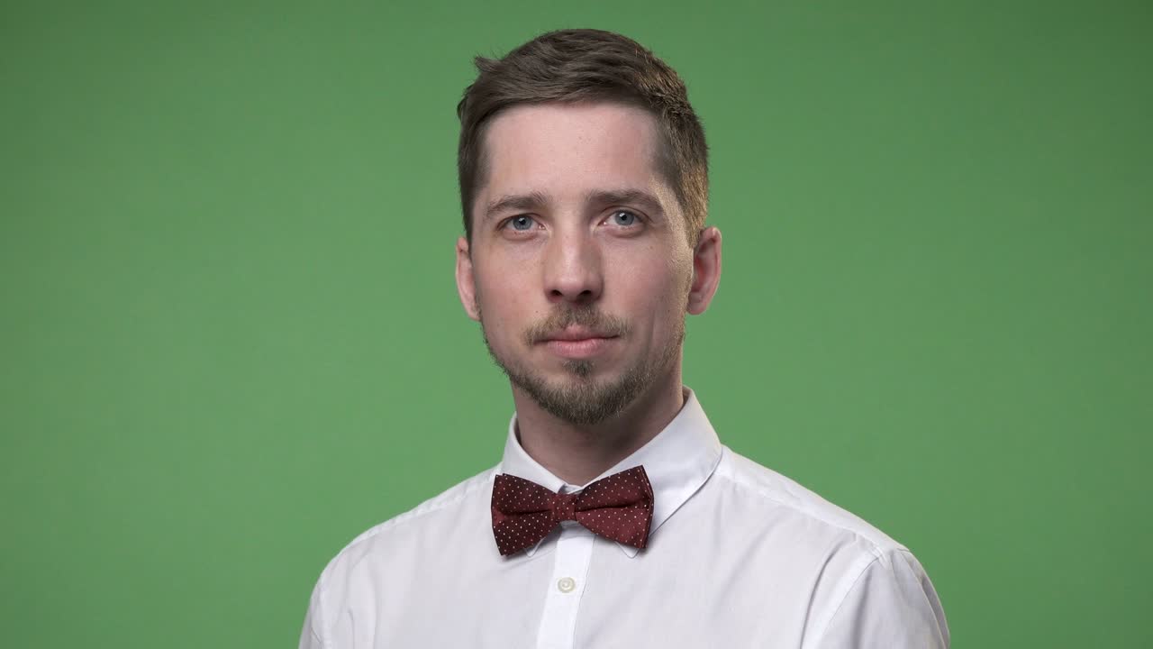 Serious young man in a white shirt with a tie butterfly turns his head, chroma key green screen background