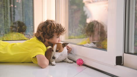 niño rubio con el pelo rizado tirado en el suelo con su perro mientras lo acaricia y mira por la ventana