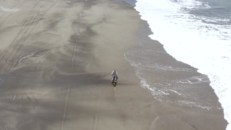 dirt bike rider fun ride on a sandy beach in bali, indonesia with calm waves breaking, top down aerial view