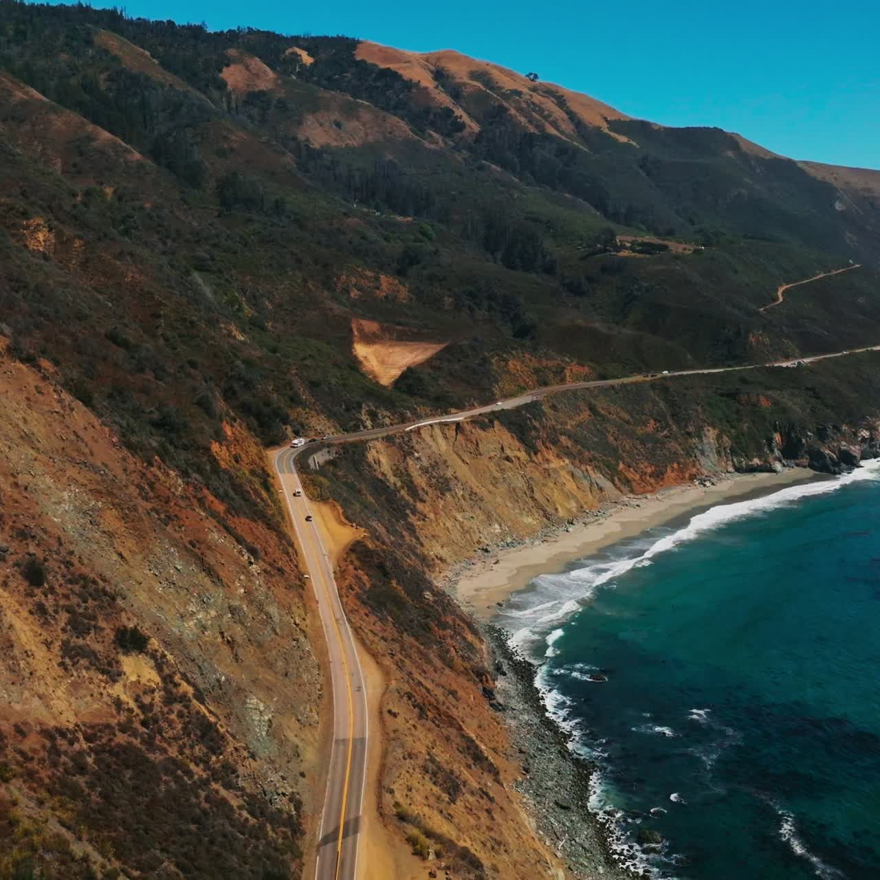 Road along the sloping mountainous coast of Pacific in California. Amazing scenery of the shoreline on sunny hot day. Top view