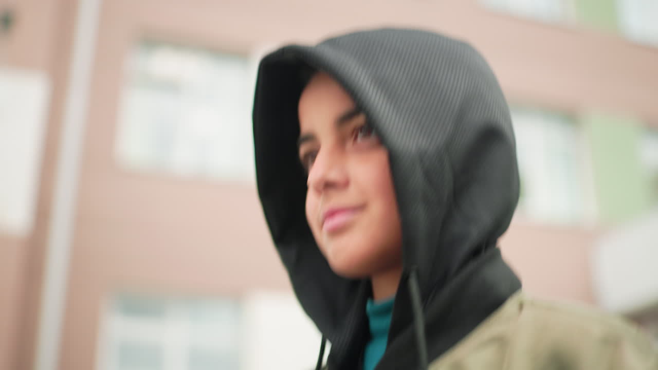 primer plano de un chico con sudadera y una sonrisa cálida caminando al aire libre cerca del edificio de la escuela, fondo borroso con ventanas que resaltan la expresión juvenil y un ambiente académico informal con luz tenue