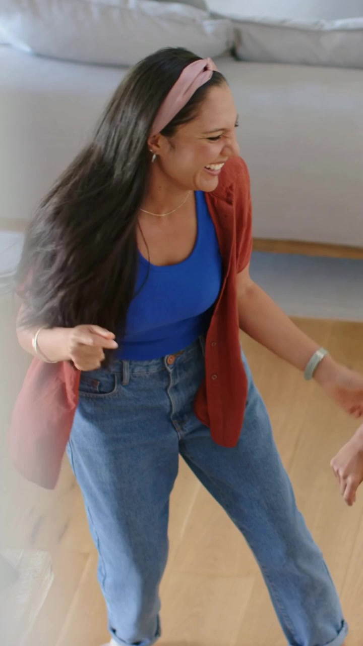 Vertical video: Dancing woman in blue tank rust shirt stepping at home, reacting to hand by sofa