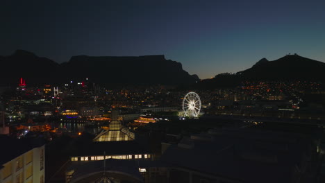 Backwards-fly-above-illuminated-large-building-of-shopping-centre.-Ferris-wheel-glowing-into-night.-Cape-Town,-South-Africa