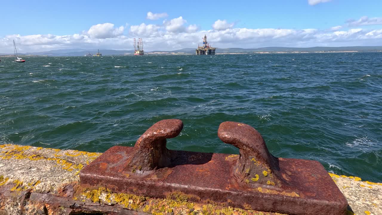 A stationary camera captures a rusty mooring cleat on a stone pier with waves, distant sailboats, and an offshore platform under bright daylight in Cromarty, Scotland