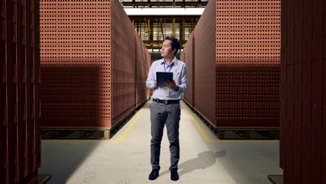 full body side view of an asian male professional worker with red brick packed in stacks are stored, observes by looking up before he come to concentrating on the tablet and keep on typing