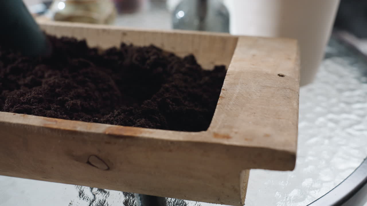 Close up of wooden planter on glass table as female hand spreads fine sand with blue plastic scoop inside planter under soft natural light indoors with blurred pot and tabletop details