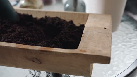 close up of wooden planter on glass table as female hand spreads fine sand with blue plastic scoop inside planter under soft natural light indoors with blurred pot and tabletop details