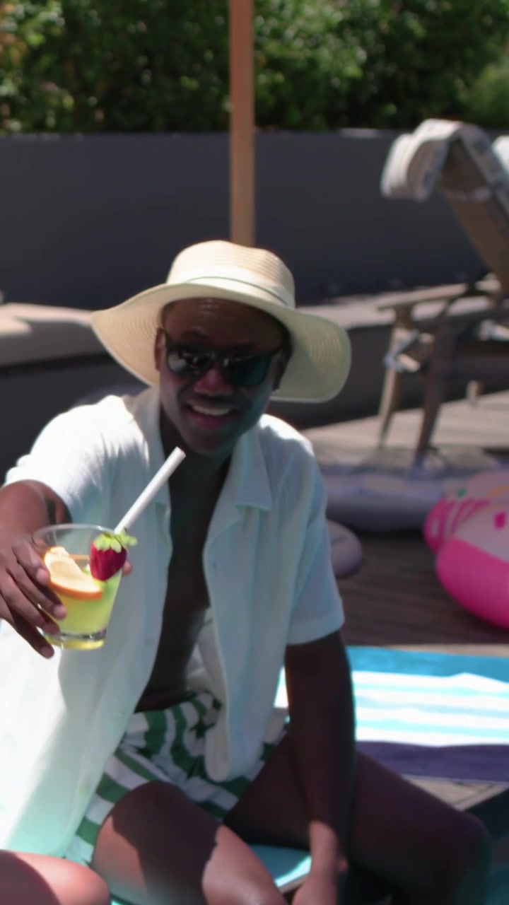 Video verticale: Telecamera che si allontana, uomo con cappello di paglia che alza un cocktail giallo a bordo piscina, condividendo un sorso.