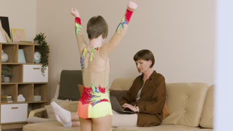 Niña-Vestida-De-Gimnasia-Practicando-Frente-A-Su-Madre,-Que-Está-Usando-Una-Laptop