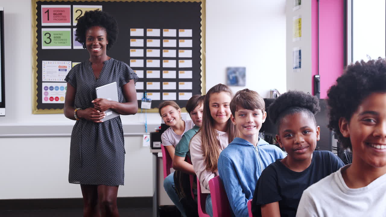 Portrait Of Female Teacher Holding Digital Tablet With Line Of High School Students Sitting By Screens In Computer Class