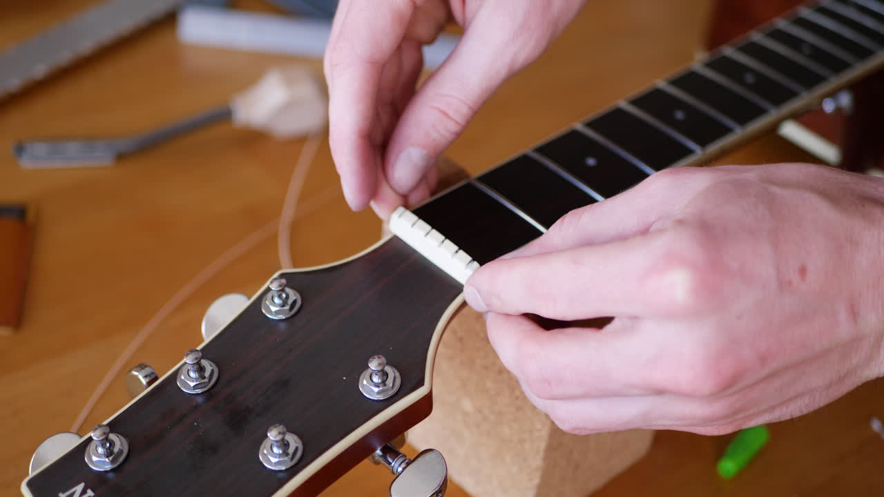 Close up hands of a luthier fitting and gluing a nut onto an acoustic guitar neck fretboard on a wood workbench with lutherie tools