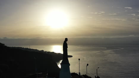 aerial view, silhouette of christ the king statue facing atlantic ocean landmark on coastline of madeira island, portugal, drone shot