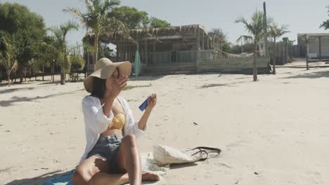 Happy-hispanic-woman-in-sunhat-using-sun-lotion-on-beach-in-the-sun,-copy-space,-slow-motion