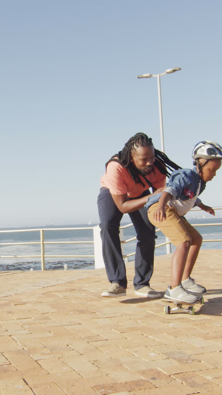 video de un feliz padre afroamericano aprendiendo a su hijo a patinar en el paseo marítimo
