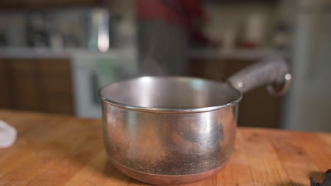 steam rising from stainless pot place on wooden table with blurry image of man at kitchen