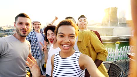 cheerful african american girl is taking selfie adjusting camera then calling her friends, posing and laughing at rooftop party. modern city is visible in background.