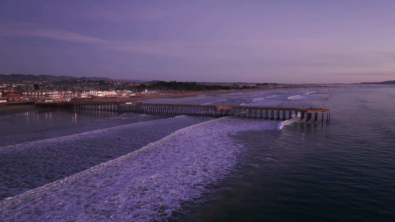 Panning to the right drone shot of the pismo beach pier at dusk with waves