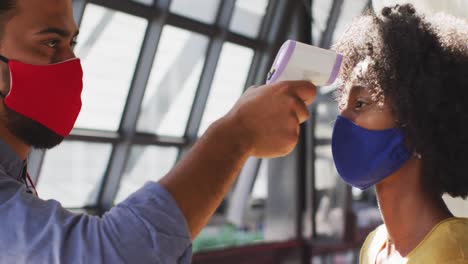 mixed race male cafe worker wearing face mask taking temperature of female coworker