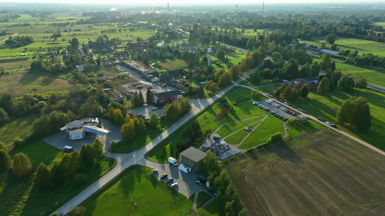 Vista aerea di Zīlāni vicino a Jēkabpils che mostra strade periferiche, aree per eventi, auto parcheggiate e i circostanti campi verdi nella calda luce della sera