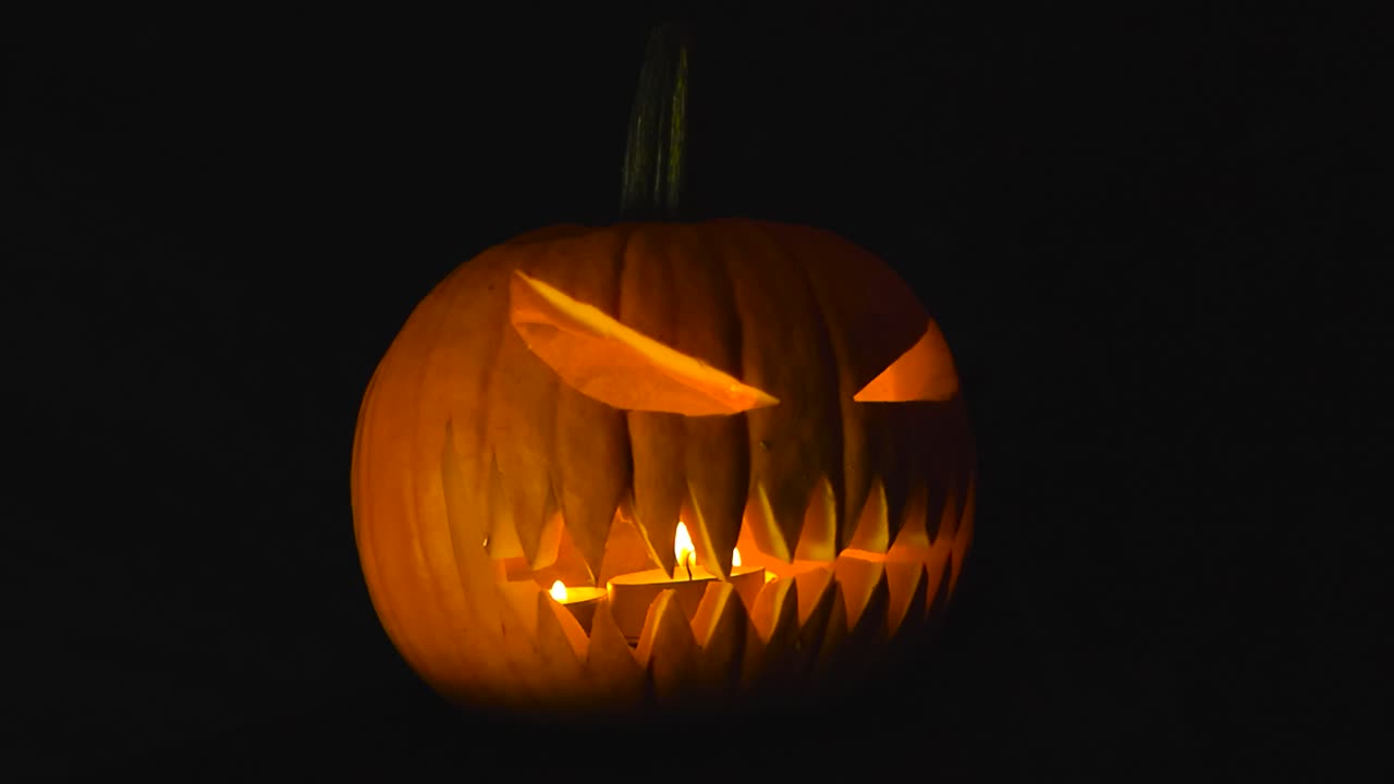 Close up video orbiting around a carved or cut orange Halloween pumpkin or Jack o Lantern that has candles inside of it burning and flickering and is in front of a black studio background. Smoke seen