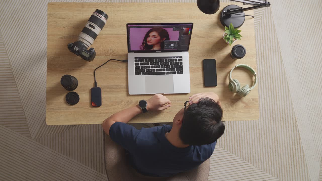 Top View Of A Male Editor Shaking His Head And Having A Headache While Sitting In The Workspace Using A Laptop Next To The Camera Editing Photo Of A Female At Home